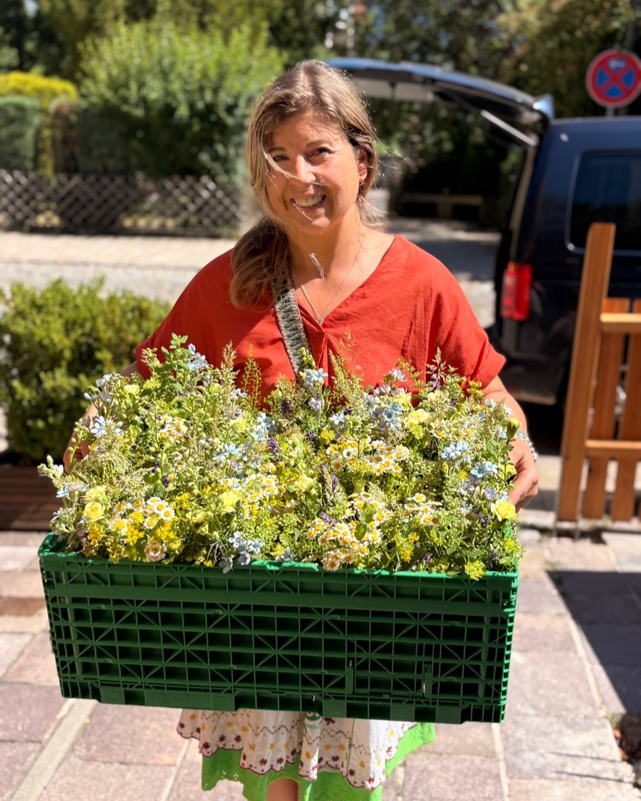 Melanie Schaper trägt lächelnd eine Kiste voller frisch geschnittener Wiesenblumen in Gelb, Weiß und Hellblau.